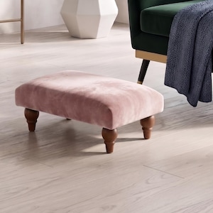 Puede incluir: Un reposapiés rectangular tapizado en suave tela de terciopelo rosa claro. La pieza cuenta con cuatro patas de madera torneada en color marrón oscuro con un diseño redondeado y bulboso que proporciona una base estable a la superficie.