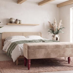 May include: A rectangular upholstered storage bench with a light beige fabric top and four dark red turned wooden legs. The bench is positioned at the foot of a bed featuring a light wood frame with a woven cane headboard and white bedding.