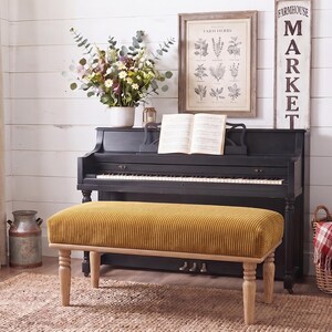 Peut inclure: Un banc rectangulaire doté d'une assise en velours côtelé jaune moutarde et de quatre pieds en bois clair au design tourné. Le banc est placé devant un piano droit noir avec un livre de musique ouvert posé sur le pupitre à partitions.
