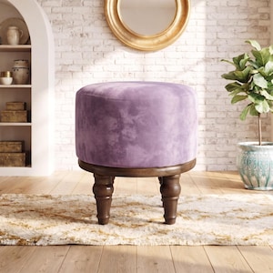 May include: A round ottoman upholstered in a soft, light purple velvet fabric. The seat is supported by four dark brown wooden legs with a turned, spindle-style design. The base of the seat features a circular wooden frame that connects the legs.
