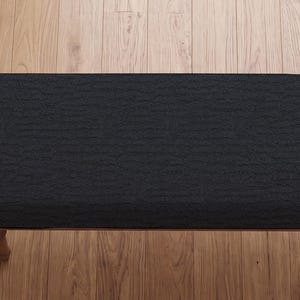 Puede incluir: Un banco rectangular con un asiento tapizado en negro con una superficie de tela texturizada y arrugada. El banco está sostenido por cuatro patas de madera de color marrón claro con un diseño torneado y redondeado, visto desde arriba.