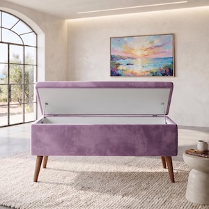 May include: A rectangular storage bench upholstered in light purple velvet fabric. The bench features a hinged lid that is open, revealing a white interior compartment. It is supported by four tapered wooden legs in a medium brown finish.