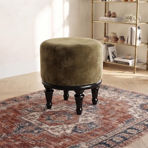 May include: A round ottoman upholstered in olive green velvet fabric. The seat features a smooth, cylindrical top and is supported by four dark brown, turned wooden legs that provide a stable base for the piece of furniture.