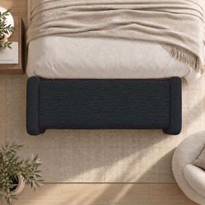 May include: A rectangular charcoal gray upholstered bench with rounded bolster ends. The bench features a textured fabric surface and is positioned horizontally in front of a bed with a light beige blanket and a white duvet cover.