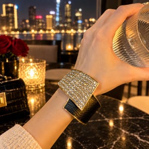 May include: A wide cuff bracelet featuring a rectangular panel covered in clear, sparkling crystals layered over a smooth, black textured leather band. The design incorporates a gold-toned metal frame that secures the two contrasting materials together.
