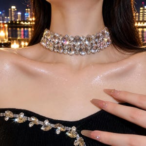 Op de afbeelding: Een brede choker-halsketting met meerdere rijen heldere, gefacetteerde, ovale en ronde kristallen op een metalen basis. De kristallen zijn gerangschikt in een dicht, gelaagd patroon dat de nek bedekt en het licht op een schitterende wijze weerkaatst.