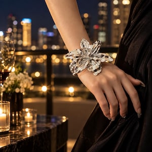 May include: A silver-toned bracelet featuring a large, dimensional flower centerpiece with metallic petals, attached to a band composed of multiple strands of small, round, grey beads that wrap around the wrist.