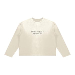Puede incluir: Una camiseta de manga larga de color crema con el texto negro "Abandon all hope, ye who enter here" impreso en una fuente de estilo gótico en el centro del pecho. La prenda tiene cuello redondo y un corte holgado y cuadrado.