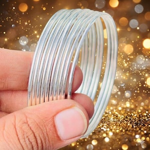 Puede incluir: Un conjunto de siete brazaletes de metal de color plateado, finos y lisos, apilados juntos. Las pulseras tienen un acabado pulido y reflectante con un diseño circular sencillo. Cada brazalete tiene un grosor uniforme y coherente.
