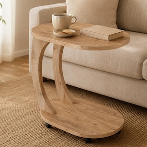 May include: A light brown wood-grain C-shaped side table with an oval top and base, featuring a curved support pillar and four small black caster wheels underneath. The table surface holds a ceramic mug and a small rectangular book.