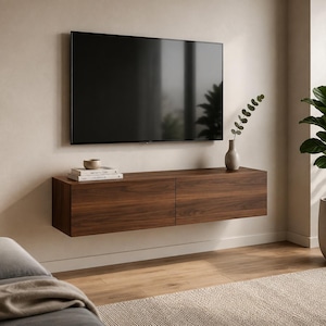 Puede incluir: Una consola multimedia rectangular montada en la pared con un acabado de grano de madera de nogal oscuro y dos puertas de armario. Un televisor de pantalla plana está montado sobre la consola, que sostiene dos libros apilados y un jarrón.