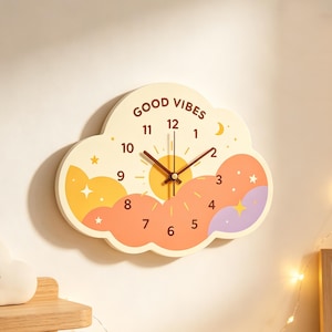 May include: A cloud-shaped wall clock with a cream-colored background featuring the text GOOD VIBES at the top. The clock face displays a yellow sun, orange and purple clouds, and small stars. It has dark brown numbers and clock hands.