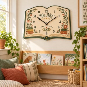 May include: A wall clock shaped like an open book with a cream-colored face, black numerals, and black hands. The text reads Mrs. Kent and Read Grow Shine. It features illustrations of stacked books, a coffee mug, and floral vines in green and red.