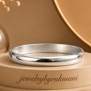 May include: A polished silver-toned bangle bracelet with a smooth, rounded surface. The bracelet is displayed on a circular tan pedestal featuring the text jewelrybyrukmani printed in a dark, elegant, cursive font on the front side.