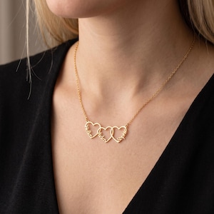 May include: A gold-toned necklace featuring three interconnected heart-shaped pendants. Each heart is engraved with a name in a cursive script, specifically the names Hellen, Aiden, and Sophie. The pendant is suspended from a delicate gold-toned chain.
