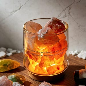 Puede incluir: Un recipiente cilíndrico de vidrio lleno de rocas de sal translúcidas de color naranja brillante, sobre una base circular de madera. El vidrio tiene un texto impreso en negro: What we should care about is not where it's from, who can shoulder the most risk will gain the most.