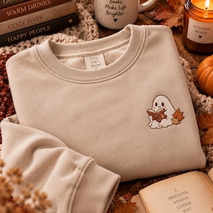 Könnte beinhalten: Ein beigefarbenes Sweatshirt mit Rundhalsausschnitt und einer Stickerei, die einen kleinen weißen Geist zeigt, der ein braunes Buch neben orangefarbenen Herbstblättern liest. Das Etikett am Innenkragen zeigt den Text Cozy Days Ahead. Der Stoff ist weich.