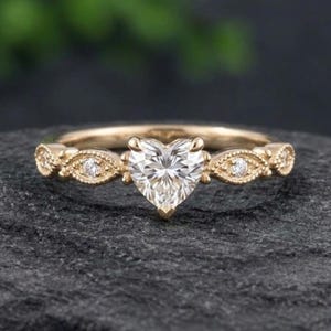 May include: A gold ring featuring a central heart-shaped clear gemstone. The band is accented with small round stones set within marquise-shaped metal frames, showcasing a detailed milgrain texture along the edges of the setting.