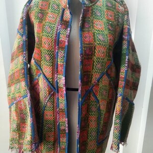 Könnte beinhalten: Eine langärmelige Jacke mit einem lebendigen, mehrfarbigen Patchwork-Muster aus grünen, orangefarbenen und rosafarbenen Quadraten. Das Kleidungsstück hat einen Stehkragen, blaue Paspeln an den Nähten und der Vorderseite sowie einen ausgefransten Saum.