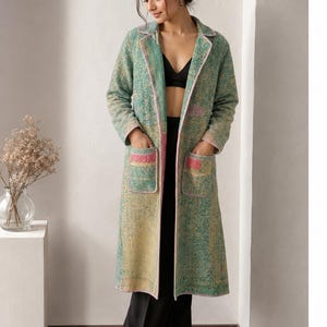 May include: A long-length coat featuring a textured, multi-coloured pattern in shades of green, yellow, and pink. The coat has a notched collar, long sleeves, and two large front patch pockets with pink trim. It is worn over a black top and trousers.
