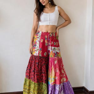 Puede incluir: Un top corto blanco sin mangas con botones en la parte delantera, combinado con pantalones palazzo anchos de varias capas con un diseño de patchwork de paneles de tela rojos, amarillos, verdes y morados con varios patrones florales y geométricos.