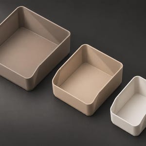 May include: Three open-top rectangular storage bins arranged by size. The largest bin is a dark taupe, the medium bin is a light beige, and the smallest bin is an off-white. Each container features a low-profile front edge and smooth walls.