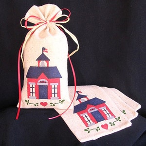 May include: Two white fabric bags with red and blue schoolhouse designs. The bags are tied with red and yellow ribbon. The schoolhouse design includes a red roof, blue walls, and white windows. The bags are decorated with green leaves and a red heart.