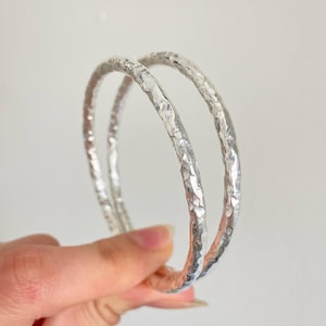 May include: Two circular silver bangles featuring a distinct hammered texture across the entire surface. The metal has a reflective, polished finish that catches the light, creating a series of small, irregular indentations along the bands.