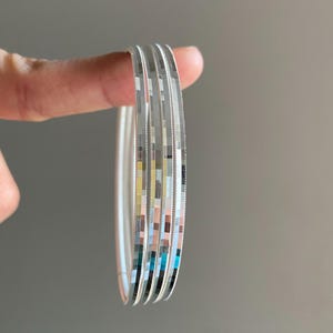 May include: A set of four silver-toned bangles featuring a reflective, multi-faceted surface with a repeating square pattern. Each bangle has a smooth, polished interior and a textured, shimmering exterior that catches the light.