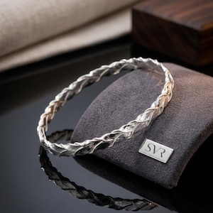 Op de afbeelding: Een zilveren armband met een doorlopend, ingewikkeld gevlochten of gedraaid metaalontwerp. Het oppervlak van het metaal heeft een gepolijste, reflecterende afwerking. Een klein rechthoekig metalen label met de inscriptie SVR is bevestigd.