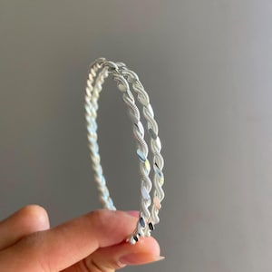 Könnte beinhalten: Ein Paar silberfarbene Armreifen mit einer gedrehten, seilartigen Textur. Die Armbänder haben eine polierte, reflektierende Oberfläche und eine kreisförmige Form. Das Design besteht aus zwei einzelnen Strängen, die zu einer Einheit verbunden sind.