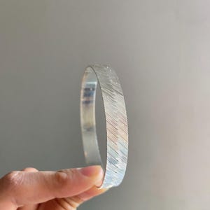 May include: A silver-toned bangle bracelet featuring a textured surface with fine, diagonal etched lines. The metal band has a polished finish and a consistent width, showcasing a metallic sheen that reflects light across its detailed, grooved pattern.