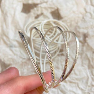 May include: A set of thin, silver-colored bangles featuring a textured herringbone pattern along the outer surface. The bracelets are circular and metallic, with a reflective finish that catches the light, showcasing a repeating chevron design.