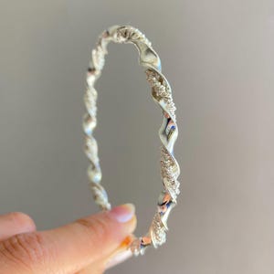 Puede incluir: Una pulsera tipo brazalete de tono plateado con un diseño trenzado que alterna secciones lisas y pulidas con segmentos texturizados y granulares. La banda metálica circular presenta un patrón continuo y repetitivo en toda su circunferencia.