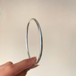 May include: A simple, circular bangle bracelet with a smooth, polished silver-toned finish. The band has a thin, rounded profile and a reflective metallic surface that catches the light, creating a clean and uniform appearance throughout the piece.
