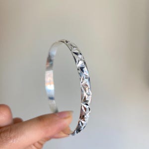 Peut inclure: Un bracelet jonc de couleur argentée présentant un motif continu de gravures facettées en forme de losange sur la surface extérieure. Le métal possède une finition polie hautement réfléchissante qui capte la lumière, créant un effet étoilé.