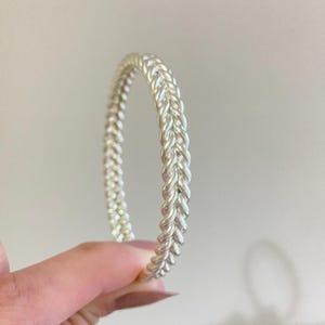 Pode incluir: Um bracelete de tom prateado com uma textura contínua trançada ou torcida semelhante a uma corda em toda a sua circunferência circular. A superfície metálica tem um acabamento polido e reflexivo que capta a luz ao longo do padrão.