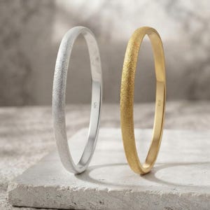 May include: Two thin, circular bangles standing upright. One bangle is silver-toned with a textured, matte finish, and the other is gold-toned with a matching textured finish. Each bangle features the small, stamped text 925 on the inner surface.