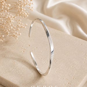Puede incluir: Una pulsera tipo brazalete de tono plateado con una superficie retorcida y reflectante, con un diseño abierto. La palabra OKROS está impresa en letras mayúsculas blancas en la parte inferior central del marco, debajo de la pulsera.