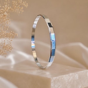 May include: A silver-toned bangle bracelet featuring a faceted, geometric design with multiple flat, reflective surfaces. The band has a consistent width and a polished metallic finish that catches light, creating alternating highlights and shadows.