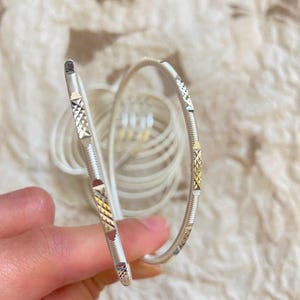 Op de afbeelding: Een dunne, ronde zilverkleurige armband met een getextureerd oppervlak met afwisselende gladde, geribbelde en diamantgeslepen secties. De metalen band heeft een reflecterende afwerking en een herhalend geometrisch patroon over de omtrek.