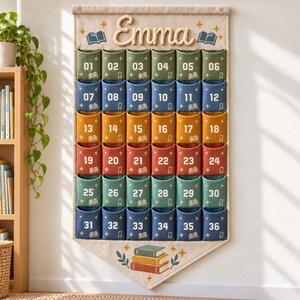 Puede incluir: Un organizador de pared de tela beige con el nombre Emma en letras de cuerda. Tiene treinta y seis bolsillos numerados en filas de colores verde, azul, dorado y rojo, decorados con pequeños iconos de libros y estrellas.