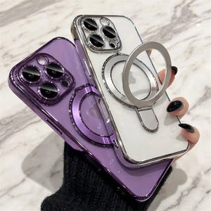 May include: Two smartphone cases with metallic edges and integrated circular magnetic rings. One case is a deep purple with a matching camera lens frame, while the other is clear with a silver camera frame and a silver magnetic ring stand.