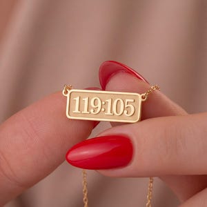 May include: A gold-toned necklace featuring a rectangular pendant with the engraved numbers 119:105. The pendant is attached to a delicate gold-toned chain, showcasing a polished finish and a clean, structured design for a classic jewelry piece.