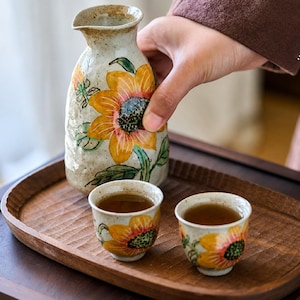 Può includere: Una caraffa per sake in ceramica e due tazzine abbinate, tutte caratterizzate da una smaltatura bianco sporco maculata con disegni di girasoli gialli dipinti a mano. La caraffa ha un collo stretto e un beccuccio, le tazze sono rotonde.