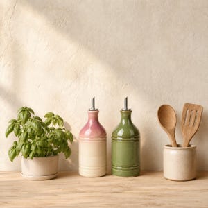 Può includere: Una pianta di basilico verde in un vaso di ceramica, due dosatori per olio in ceramica con beccucci in metallo rosa e verde, e un portautensili in ceramica contenente un cucchiaio e una spatola di legno su una superficie di legno.