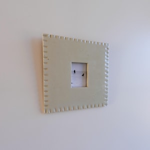 May include: A square, light beige ceramic picture frame with a textured, scalloped edge border. The frame features a central rectangular opening containing a small photograph of two dark figures standing on a vast, light-coloured, open landscape.