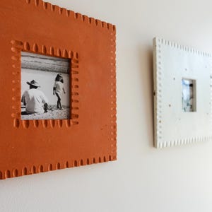 May include: A square terracotta-coloured picture frame with a textured, notched border containing a black and white photograph of a person in a hat and a child. A second, smaller white square frame with a similar notched border is visible nearby.