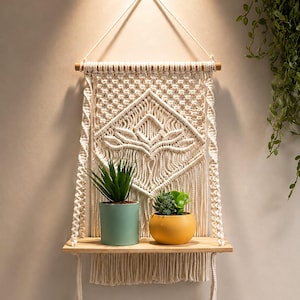 May include: A cream-coloured macrame wall hanging featuring a central lotus-like knot pattern, suspended from a wooden dowel with a flat wooden shelf. Two small potted plants, one in a light blue pot and one in a yellow pot, sit on the shelf.