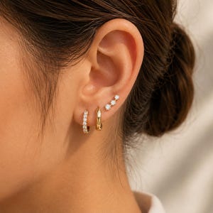 May include: A set of three gold-toned earrings worn in a single ear. The set includes a small hoop earring with embedded clear stones, a plain polished gold hoop earring, and a small stud earring featuring three aligned clear stones.